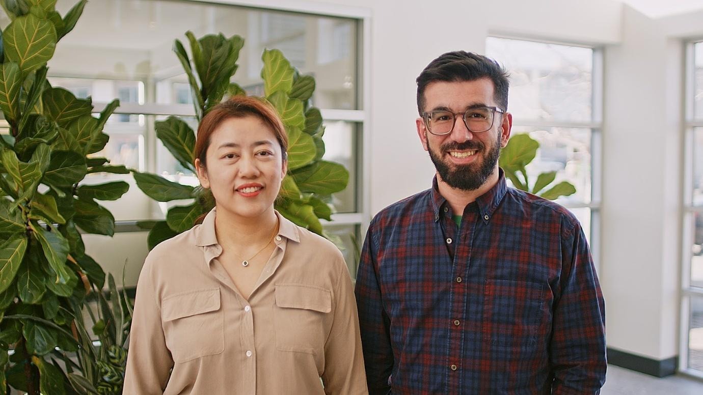 Two people are standing in an office space with potted plants and large windows behind them.
AI-generated content may be incorrect.